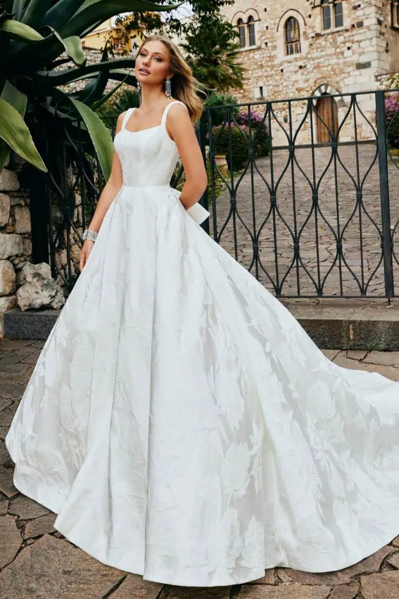 Sima, egyszerű esküvői ruha brokát anyagból. Bodyja szolíd kivágású, széles vállpántú, a ruha hátuját pedig masni díszíti. Style: Veni Infantino "52075"